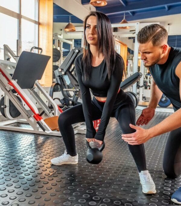 Personal trainer coaching female bodybuilder making plie squat with dumbbell